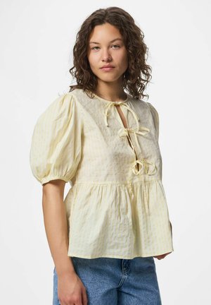 Kobieta o kręconych włosach, ubrana w bladożółtą bluzkę z bufkami i trzema kokardkami z przodu, połączoną z niebieskimi dżinsami, zwrócona przodem.