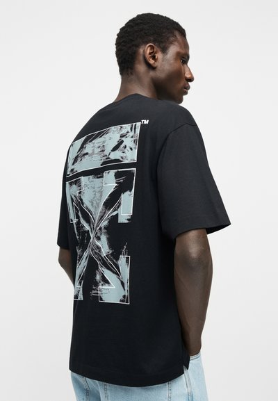 Άνδρας που φοράει μαύρο φαρδύ μπλουζάκι με μεγάλο άσπρο και γκρι αφηρημένο σχέδιο βέλους στην πλάτη, συνδυασμένο με ανοιχτό μπλε τζιν παντελόνι.