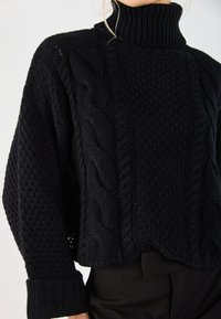 Pull à col montant en maille noire avec des motifs texturés, des détails en tricot torsadé et des poignets côtelés. Coupe loose avec un col haut.