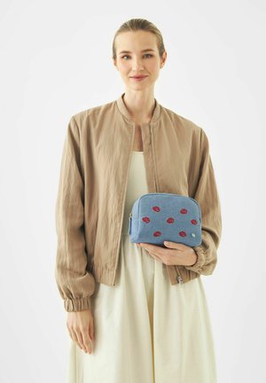 Woman with light skin and blonde hair in beige jacket and cream dress holding blue pouch with red lip print patterns.