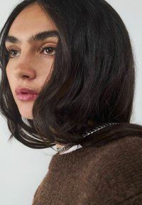 Brown textured sweater with a round neckline, paired with a silver chain necklace. Dark wavy hair frames the face, emphasizing warm skin tones.