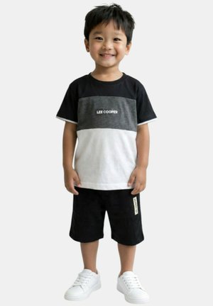 Jeune garçon souriant debout, portant un t-shirt Lee Cooper noir, gris et blanc, un short noir avec une étiquette, et des baskets blanches.
