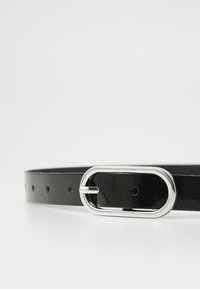 Svart lakkert lærbelte med en skinnende sølvoval spenne, som har flere justeringshull for størrelsesjustering. Glatt tekstur, elegant design.