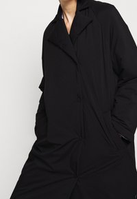 Schwarzer oversized Mantel mit einem Reverskragen, ausgestattet mit einem Knopfverschluss und strukturiertem Stoff. Taschen sind in das Design integriert.