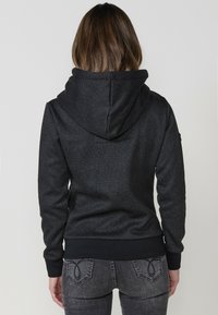 Felpa nera con un motivo testurizzato, dotata di un grande cappuccio, polsini a coste e orlo elastico. Il tessuto appare morbido e caldo.