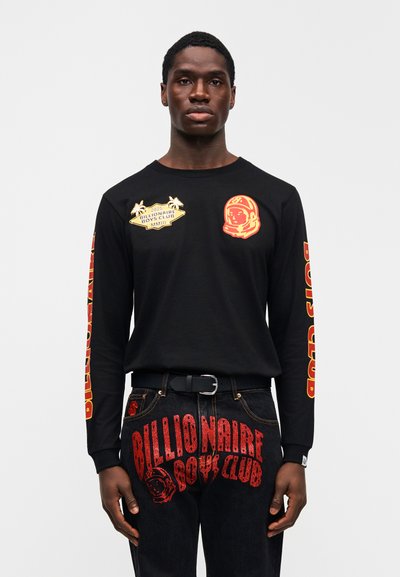 Μαύρο μακρυμάνικο πουκάμισο με κόκκινα και κίτρινα γραφικά σχέδια, που περιλαμβάνουν έναν αστροναύτη και κείμενο, συνδυασμένο με μαύρα τζιν που παρουσιάζουν παρόμοια σήμανση.