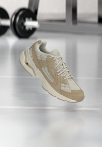 Beige atletska cipela s mrežastim i zamščenim materijalima, s teksturiranim dizajnom, vezicama i zaobljenim vrhom. Prikazana na glatkoj površini.