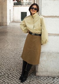 Šviesiai rusvas oversized marškinis su aukštu apykakle, derinamas su rudais zomšiniais sijonais ir juodais iki kelių siekiančiais batais. Juodas diržas su dekoratyvine sagtimi.