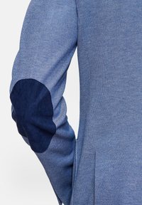 Modrá tkaná bunda se strukturovanou látkou a kontrastními tmavě modrými podložkami na loktech. Design je upraven s přiléhavým střihem.