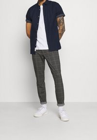 Marinblå kortärmad skjorta med knapp, vit T-shirt med rund hals under, rutiga slim-fit byxor och vita sneakers. Tatuering synlig på armen.