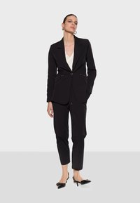 Black tailored suit featuring a fitted blazer with gold hardware accents and slim trousers. Worn over a white top, paired with black heels.