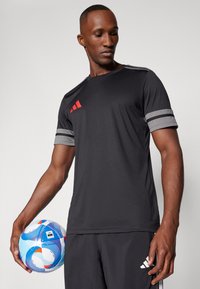Schwarzes Sport-T-Shirt mit grauen gestreiften Ärmeln, das drei rote diagonale Streifen auf der Brust hat, kombiniert mit schwarzen Shorts und einem mehrfarbigen Fußball.