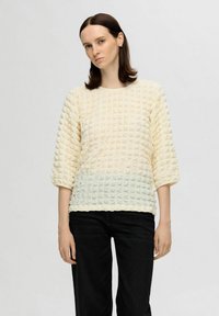 Krämfärgad jumper med textur och rutmönster, trekvartslånga ärmar och rund halsringning, i kombination med svarta byxor.