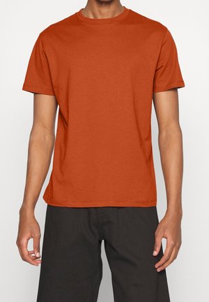 Homme portant un t-shirt uni à manches courtes orange brûlé et un pantalon noir, debout contre un fond blanc avec les bras détendus.