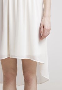 Witte chiffon jurk met een hoge-lage zoom. Zachte, lichte stof valt soepel. Hand geplaatst nabij de zoom, waarbij de vorm en het ontwerp worden getoond.