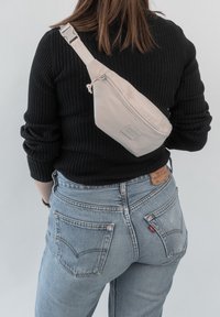 Osoba má na sobě světle modré džíny a černý žebrovaný svetr s béžovou křížovou kabelkou nošenou přes záda.