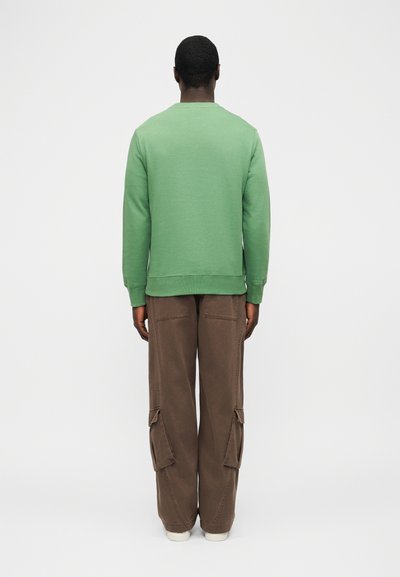 Άνδρας που στέκεται με την πλάτη γυρισμένη, φοράει πράσινο φούτερ με μακριά μανίκια και χαλαρά καφέ cargo παντελόνια με λευκά παπούτσια, σε λευκό φόντο.