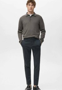 Młody mężczyzna ubrany w szarą, długorękawową koszulkę polo, ciemne zwężane spodnie, czarne skórzane loafersy, stojący z rękami w kieszeniach na białym tle.