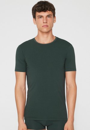 Joven con el cabello corto y rizado lleva una camiseta ajustada de manga corta verde oscuro y ropa interior a juego, de pie sobre un fondo blanco.