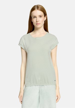 Femme aux cheveux roux lisses portant un haut texturé gris clair à manches courtes et un pantalon ample gris clair, debout contre un fond uni.