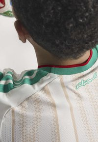 Weißes Trikot mit grünen Akzenten und rotem Kragen, das ein geometrisches Muster und strukturierter Stoff aufweist. Text in Arabisch erscheint auf dem oberen Rücken.