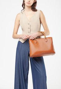 Borsa in pelle marrone con texture liscia, forma rettangolare e manico accentuato in bambù. Indossata con un top beige e pantaloni a gamba larga blu.