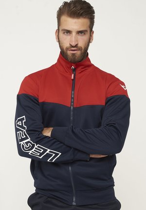 Uomo con barba che indossa una giacca con zip rossa e blu navy con la scritta "LEGEA" sulla manica sinistra, braccia incrociate, guarda dritto davanti a sé.