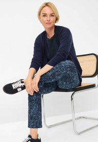 Marineblauwe cardigan, donkerblauwe gemusterde broek en zwarte sneakers met witte veters. Zittend op een stoel met een rieten zitting, handen rustend op de knieën.