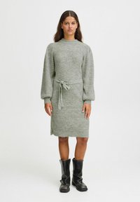 Robe en tricot en doux tissu vert clair, avec de longues manches ballons, une ceinture à la taille et des fentes sur les côtés. Assortie à des bottes noires hautes.