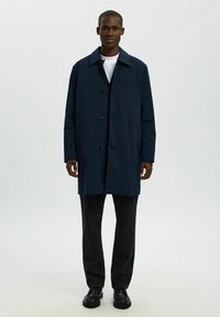 Marineblauer Mantel mit Kragen, vorderen Knöpfen und zwei Seitentaschen. Getragen über einem weißen Hemd und kombiniert mit dunkelgrauen Hosen und schwarzen Schuhen.