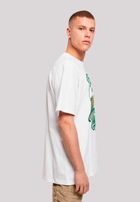 White cotton t-shirt with short sleeves, featuring green and gold graphic design on the front. Model stands sideways, wearing tan pants.
