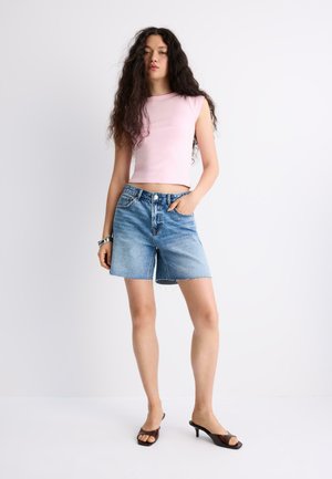 Mladá žena s dlhými kučeravými vlasmi, oblečená v ružovom bezrukávovom crop tope, riflových šortkách a čiernych sandáloch na opätku, stojaca s rukou v vrecku.