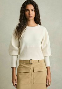 Weißer Pullover mit strukturiertem Oberteil und Puffärmeln, kombiniert mit einem beigen Rock mit zwei Vorder-Taschen und einem Gürtel.
