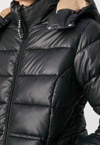 Černá prošívaná péřová bunda s lesklým vzhledem. Má vysoký límec, zapínání na zip a hnědé detaily na kapuci. Strukturální povrch.