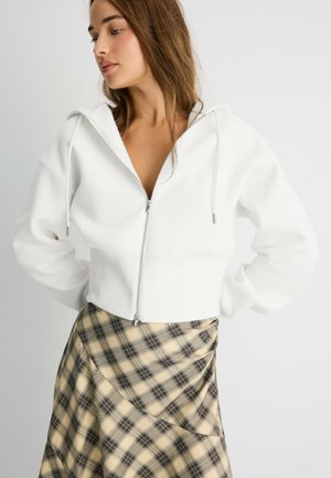 Jeune femme portant un sweat à capuche blanc zippé et une jupe à carreaux beige et noire, debout devant un fond clair uni.