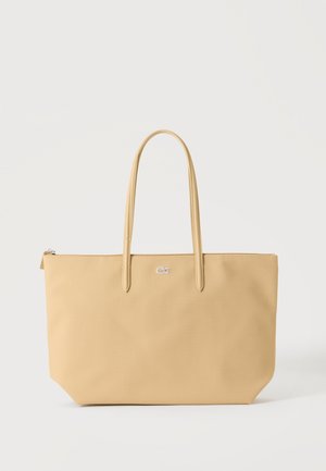 Beige rektangulær tote-taske med dobbelte håndtag og et lille sølvlogo foran i midten, sat mod en ensfarvet lys baggrund.