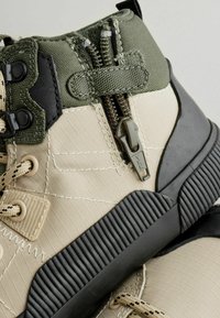 Stiefel mit einer Oberseite aus Beige und Olivgrün, mit einem Reißverschluss, strukturiertem Stoff und einer schwarzen Gummisohle. Dicke Schnürsenkel und verstärkte Nähte sind sichtbar.