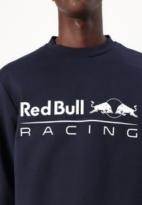 Tamno plavi dukserica od pamuka s okruglim izrezom, koja sadrži bijeli natpis "Red Bull Racing" i dvije bijele grafike bika.