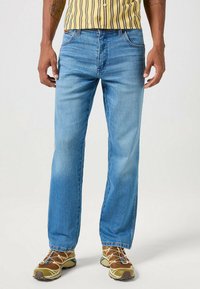 Jeans in denim azzurro chiaro con un design a gamba dritta, dotati di cinque tasche e un lieve sbiadimento. Abbinati a sneakers marroni e una camicia a righe gialla.
