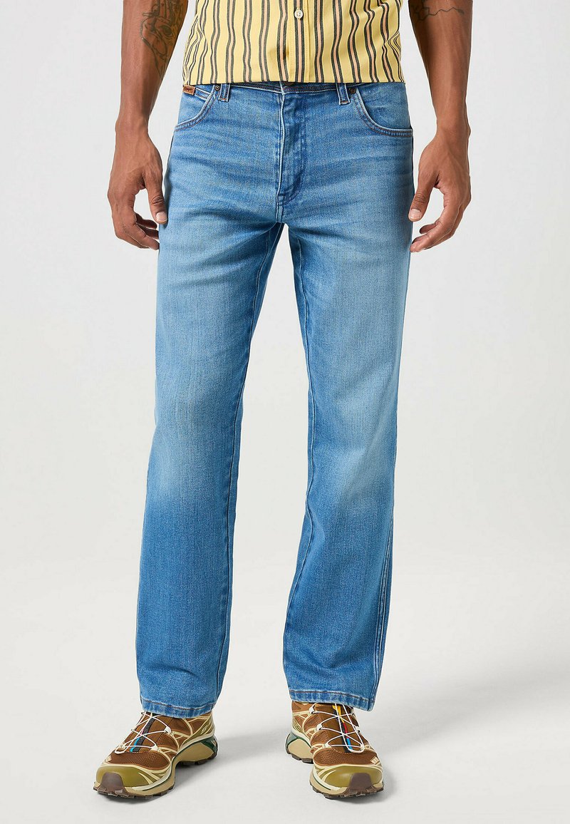 Jeans in denim azzurro chiaro con un design a gamba dritta, dotati di cinque tasche e un lieve sbiadimento. Abbinati a sneakers marroni e una camicia a righe gialla.