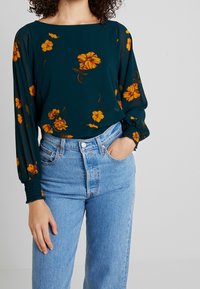 Blouse fleurie vert foncé avec des fleurs jaunes, manches longues et poignets froncés, associée à un jean taille haute bleu clair.