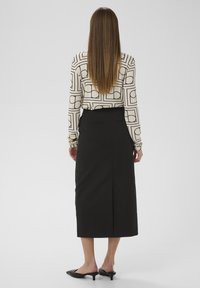 Langärmelige Bluse mit geometrischem Schwarz-Beige-Muster, kombiniert mit einem schwarzen Midirock und schwarzen spitz zulaufenden Pumps.