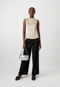 Mulher vestindo top bege sem mangas, calças pretas de perna larga, sandálias pretas de salto alto, segurando uma pequena bolsa prateada com alça de corrente.