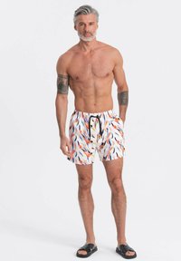Homme torse nu aux cheveux gris et tatouages, debout en short de bain blanc avec des motifs orange et bleus, et des sandales noires, les mains dans les poches.