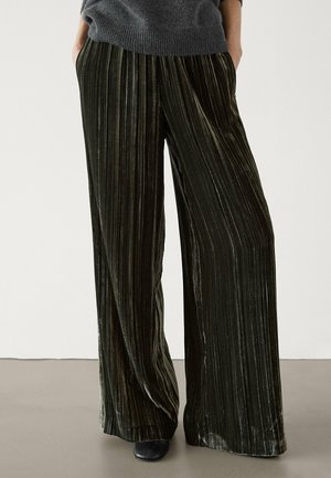 Person wearing high-waisted, wide-leg, dark green velvet pants with vertical pleats, paired with a gray sweater and black shoes.