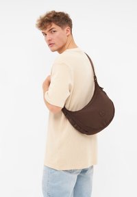 Brun nylonaxelväska med en böjd design, texturerade accenter och ett logoflik på framsidan, bärs som en crossbody, ihop med en beige skjorta och ljusa jeans.