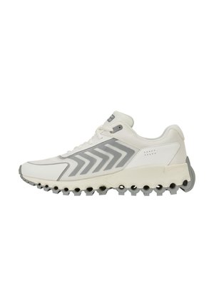 Chaussure de sport basse blanche et grise avec motif chevron sur la partie en mesh latérale et découpes circulaires distinctives sur la semelle pour l’adhérence.