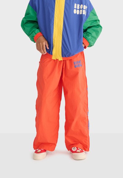 Enfant portant une veste à blocs de couleur avec une fermeture éclair jaune, un pantalon orange et des baskets rouges ornées de motifs marguerites, debout les mains dans les poches.