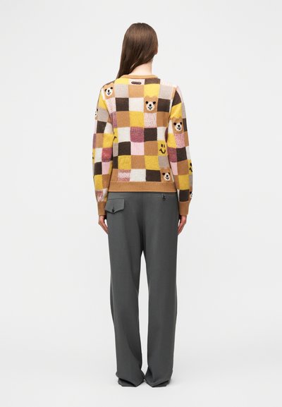 Femme aux longs cheveux bruns portant un pantalon gris et un pull à carreaux multicolore avec des visages d'ours et des motifs souriants, debout de dos.