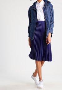 Giacca di jeans, camicia bianca con rouches, gonna blu navy plissettata e sneakers bianche. Focalizzati su stratificazione informale e contrasti di texture.